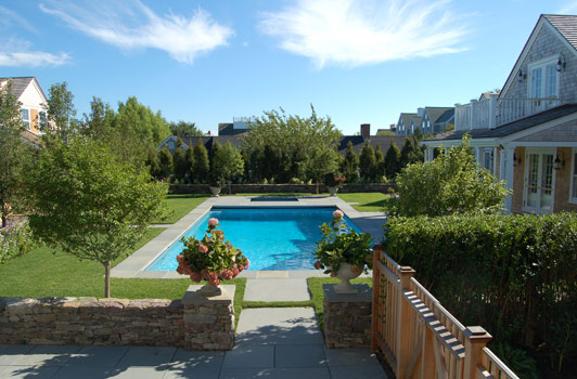 title:View of the pool descending from the main house deck, traversing the grilling/eating area. Planters flank the pool entrance and provide an opportunity for seasonal accents.